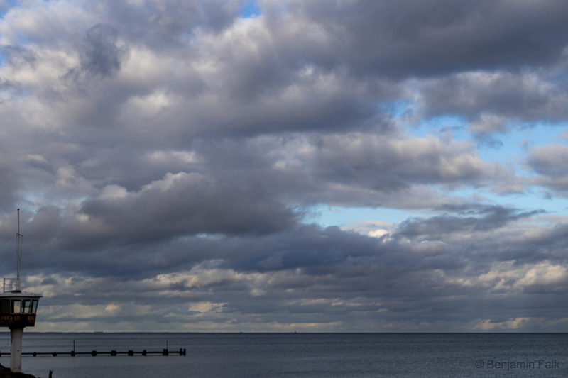 Meer unter bewölktem Himmel mit starken Kontrasten zwischen den Wolken und den Wolkenlücken. Am unteren linken Bildrand ist ein leerer DLRD Aussichtsturm mit Blick auf das Meer zu sehen.