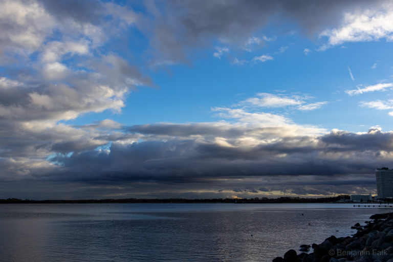 Meer, von der Küste aus fotografiert. Am linken Bildrand ist eine Steinküste, sowie am Horizont ein hotelturm zu sehen. De robere Teil des Himmels ist strhaelend blau, während vor dem utneren teil des Himmels ein Wolkenband liegt.