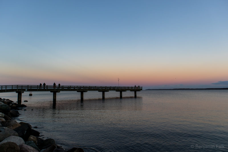 Meerbrücke von der Seite vom felsigen ufer aus aufgenommen, im Sonnenuntergang vor einem leicht rsa Himmel im Sonnenuntergang. Die Brücke spiegelt sich vor dem horizont im Wasser, auf der Brückle sind die Umrisse ovn Menschen zu sehen.