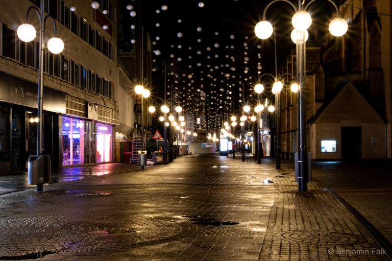Kölner Fußgängerzone aufgenommen als Langzeitbelichtung bei Nacht. Die Einkaufstraße ist menschenleer und bleiuchtet von weißen Kugelleuchten. Über der Straße hängen weitere weiße Bälle die bewegungsunscharf sind und der Boden der Straße ist übersäht von Pfützen.