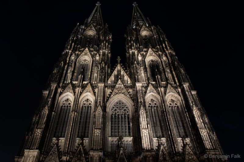Frontseite des Kölner Doms bei Nacht, aufgenommen oberhalb des Hauptportals frontal von unten mit Blick nch oben. Der Dom ist weiß angestrahlt und die gothsischen Elemente heben sich dunkel von den Weißen Flächen und Bögen ab.