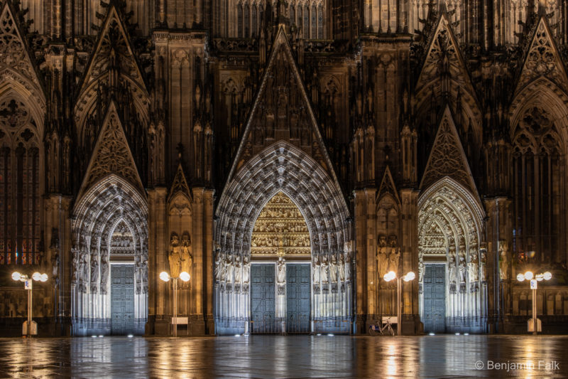 Hauptportal des Kölner Doms, bestehend aus drei Eingängen bei Nacht. Aufgenommen Frontal von vorne. Das Portal ist beleuchtet und der Platz vor dem Eingang ist regennass und die beleuchteten Bögen spiegeln sich teilweise auf dem nassen Boden.